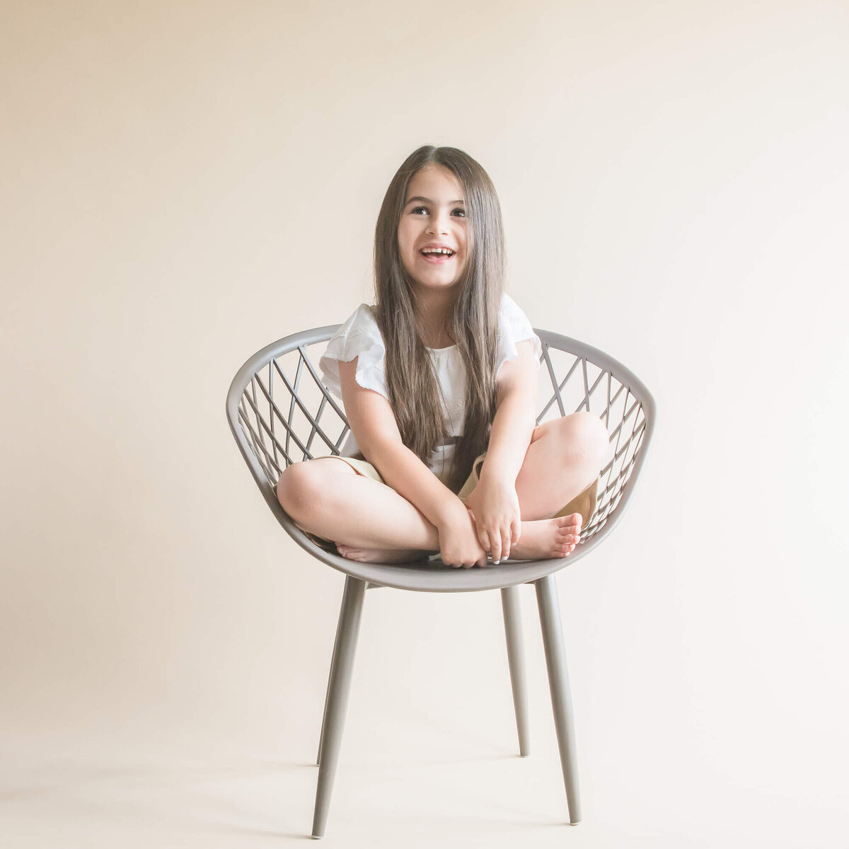 studio portrait of a young girl sitting criss cross applesauce on a grey circular chair with a cream background