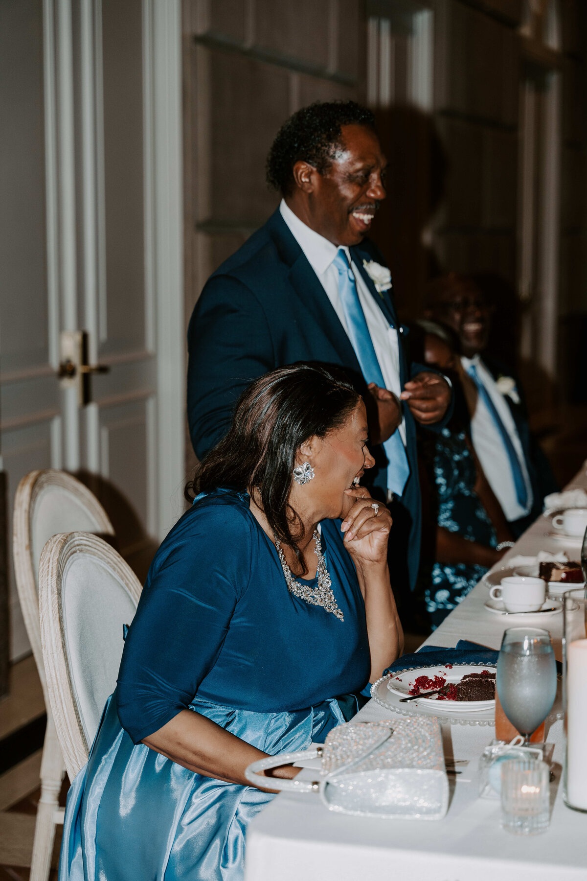 A man in a suit gives a joyful speech at a formal dinner, smiling widely. A woman in a blue dress, seated, listens and laughs, creating a warm, celebratory atmosphere.