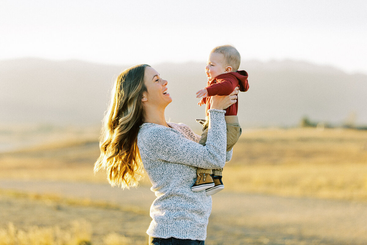 a mom twirls her baby boy in the air in front of sun setting over mountains
