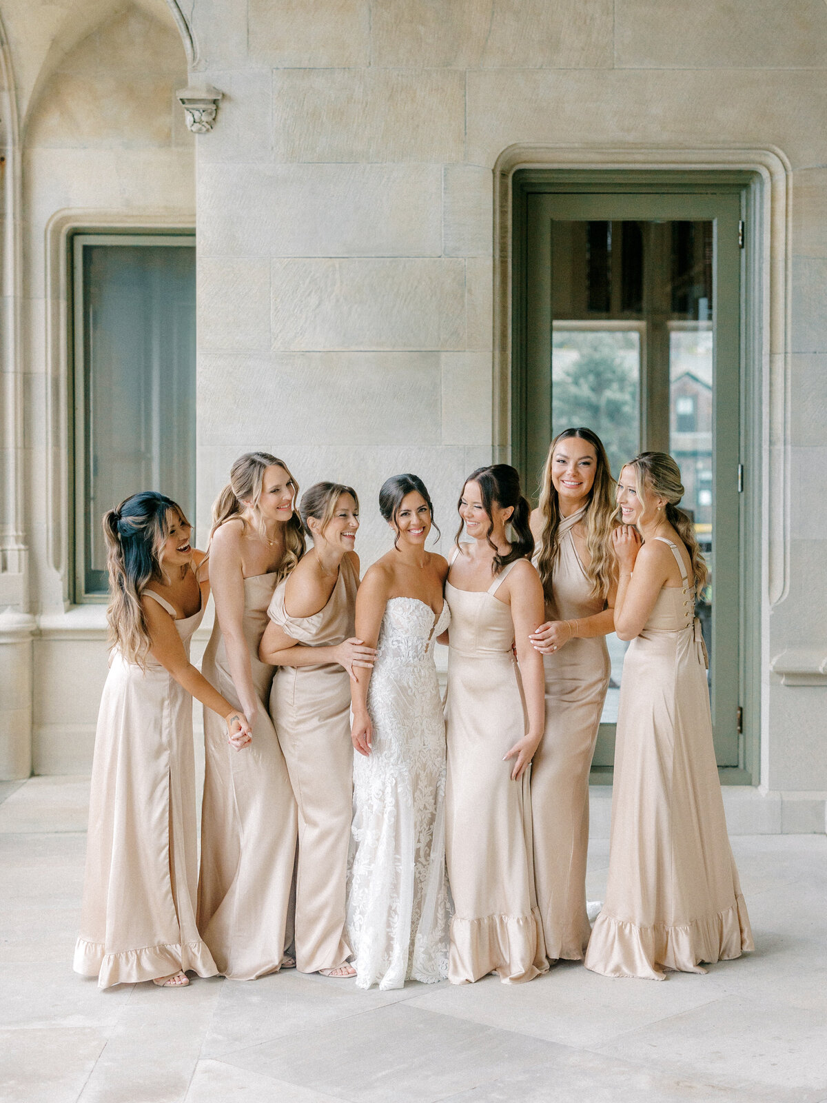 Bride with her bridesmaids in champagne colored dresses all smiling at each other in front of a stone wall