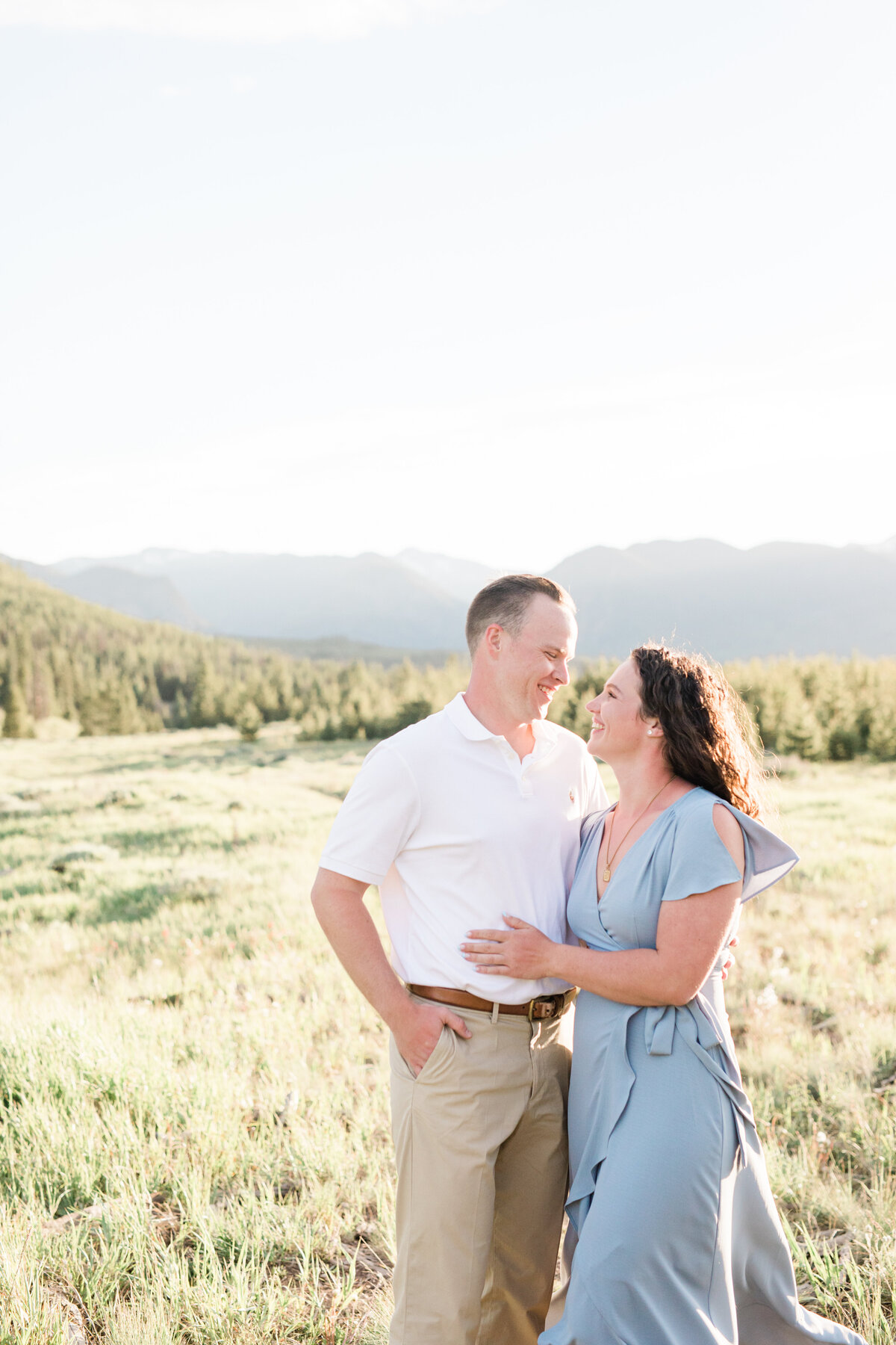 A couple in warm summer attire hold each other in a green grassy field in the ten mile range