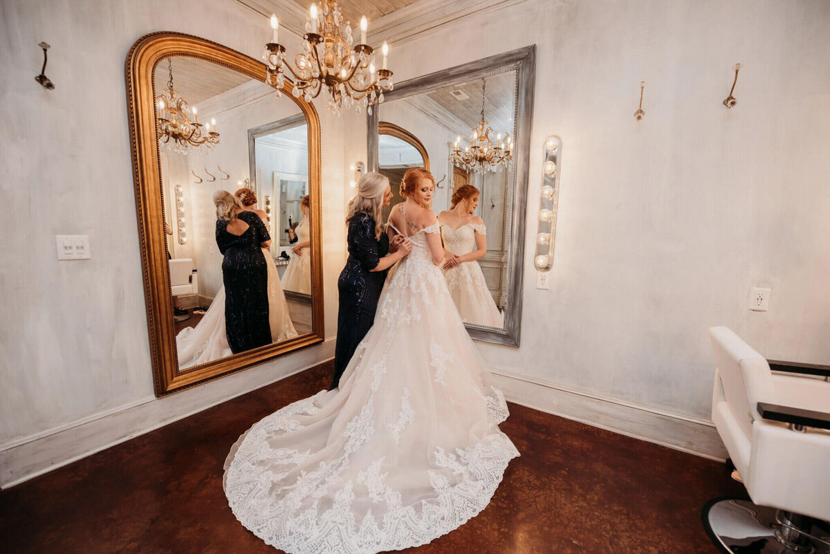 Photo of a bride getting her dress on with the help of her mother inside of a white bridal suite