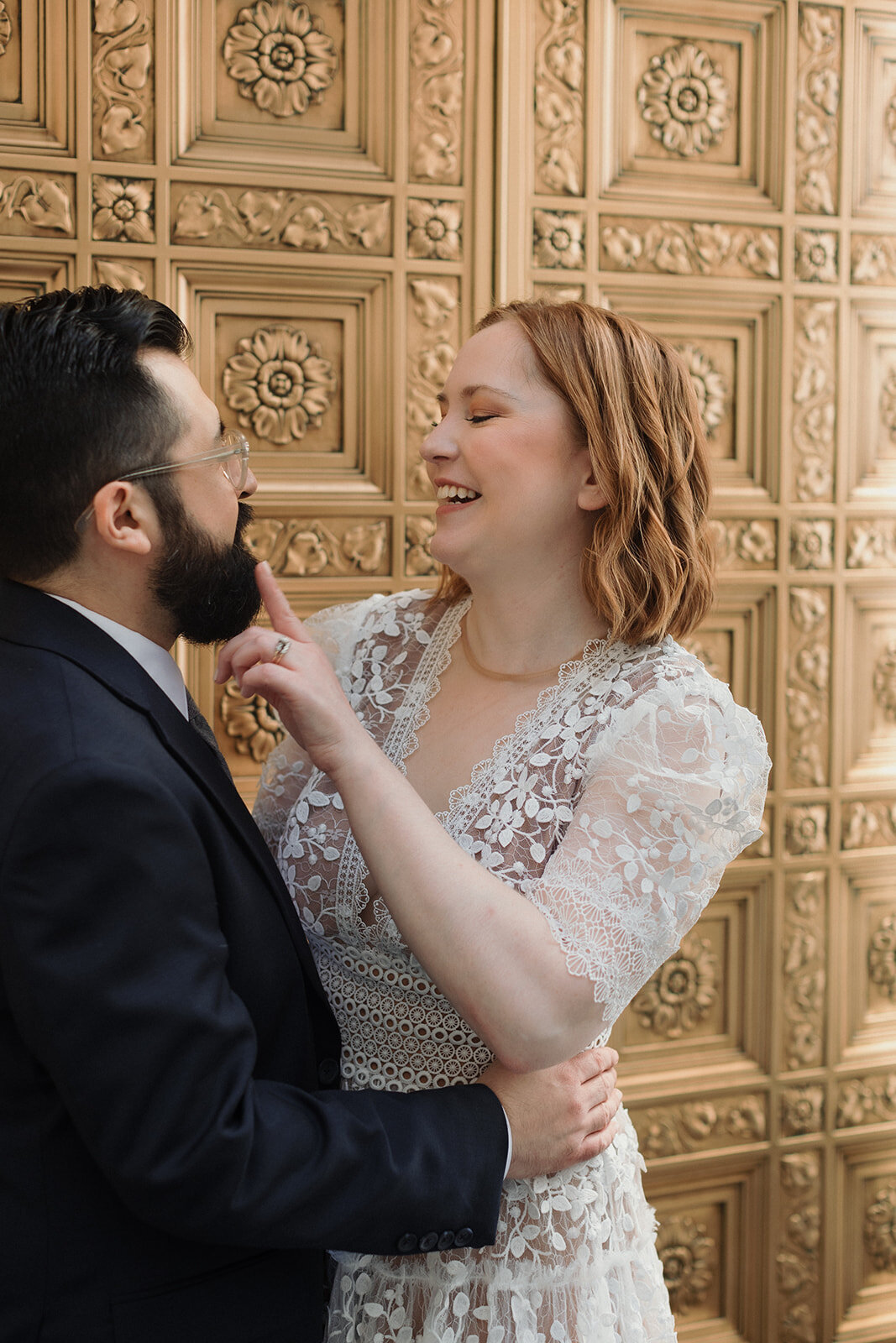 Elopement couple embraces and laughs, bride is touching the grooms face. Large gold brass doors are behind them.