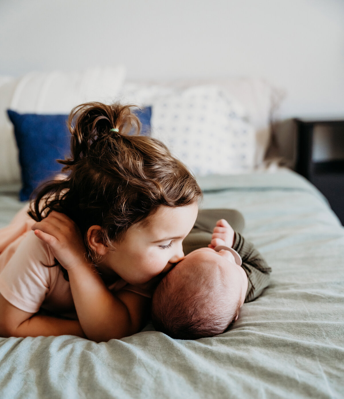 Newborn Photographer, a baby lays on the bed and older sister gives him a kiss