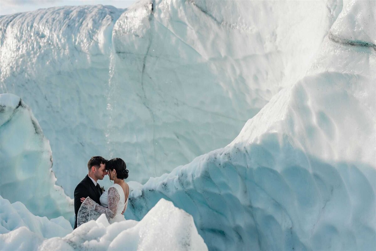 luxury elopement photographer image taken of a couple embracing while nestled in the the layers of a glacier wall in their luxury elopement in Alaska.  they are standing, with glacier ice in front of them, covering all but their upper bodies, ice in a etched wall behind them, with a third wall of ice at the very back, with ridges, and sun sheen on the ice, and a small flow of water falling over the lip of the ice and into the space behind them.  their head perfectly frame the falling water behind them.  The glacier icewalls are so big they fill the frame and the couple looks very small in the lower left corner