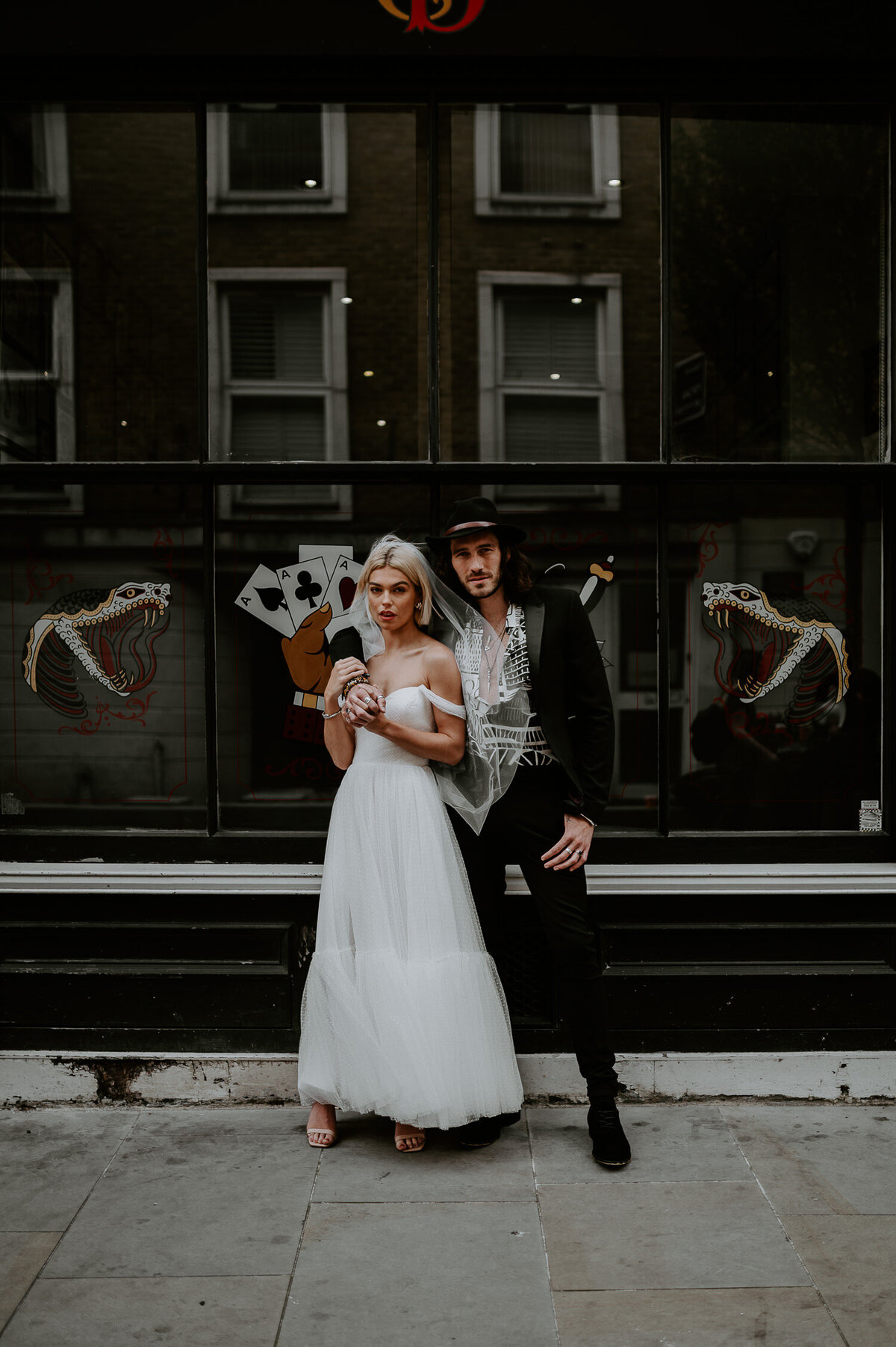 An alternative wedding couple stand outside a tattoo shop in Shoreditch London. The bride is wearing a shoulder cut wedding dress and the groom is has long hair and is wearing a casual outfit with a hat.