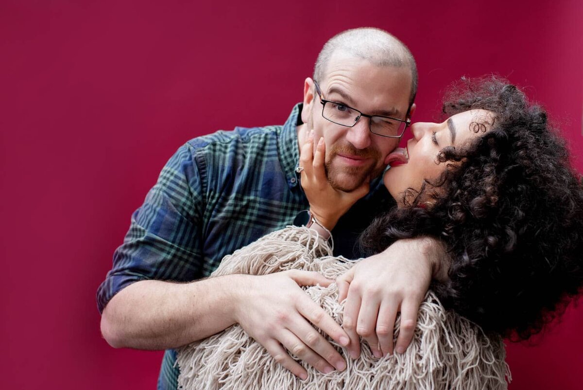 a fun woman with curly hair wearing a shaggy sweater licks her fiance's face