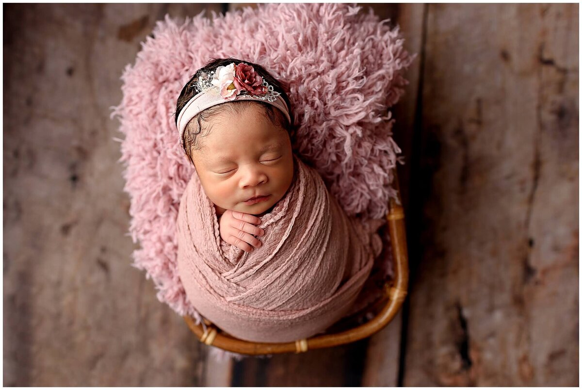 A newborn baby girl wearing a beautiful pink floral headband peacefully sleeping in a basket.
