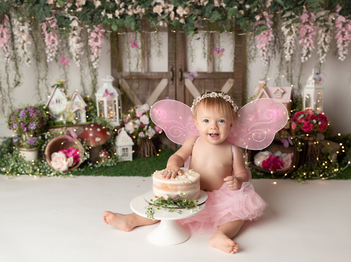 Garden Fairy Themed cake smash at West Palm Beach and Delray Beach, FL photography studio. Baby girl is wearing a pink tulle skirt and fairy wings with one hand on top of the camera. In the background, there are miniature houses, flowers, fairy lights, and mushrooms.