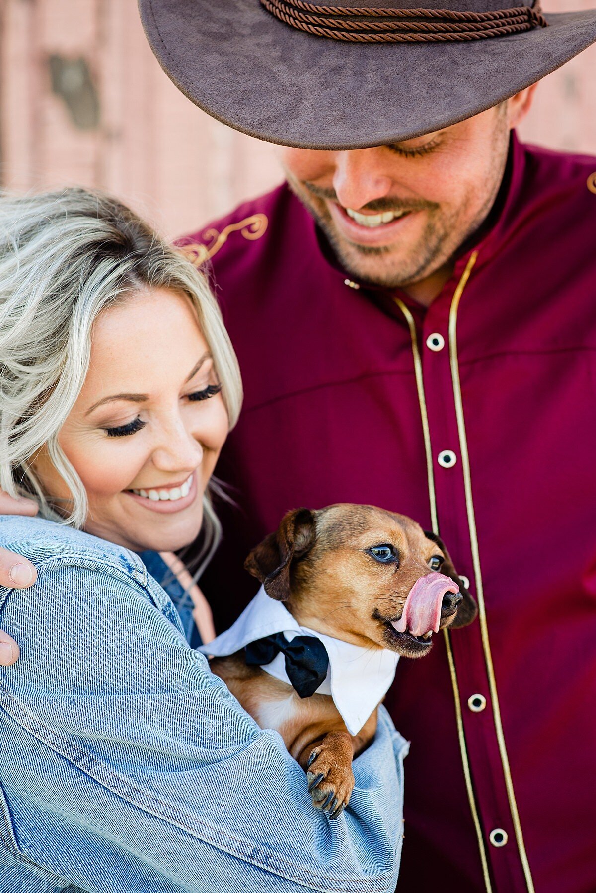The groom wearing a maroon western shirt and brown cowboy hat smiles down at the bride and their dog. The bride is wearing a denim jacket as she leans into the groom's embrace. She is holding a small dog who is wearing a white shirt collar with a small black bowtie.