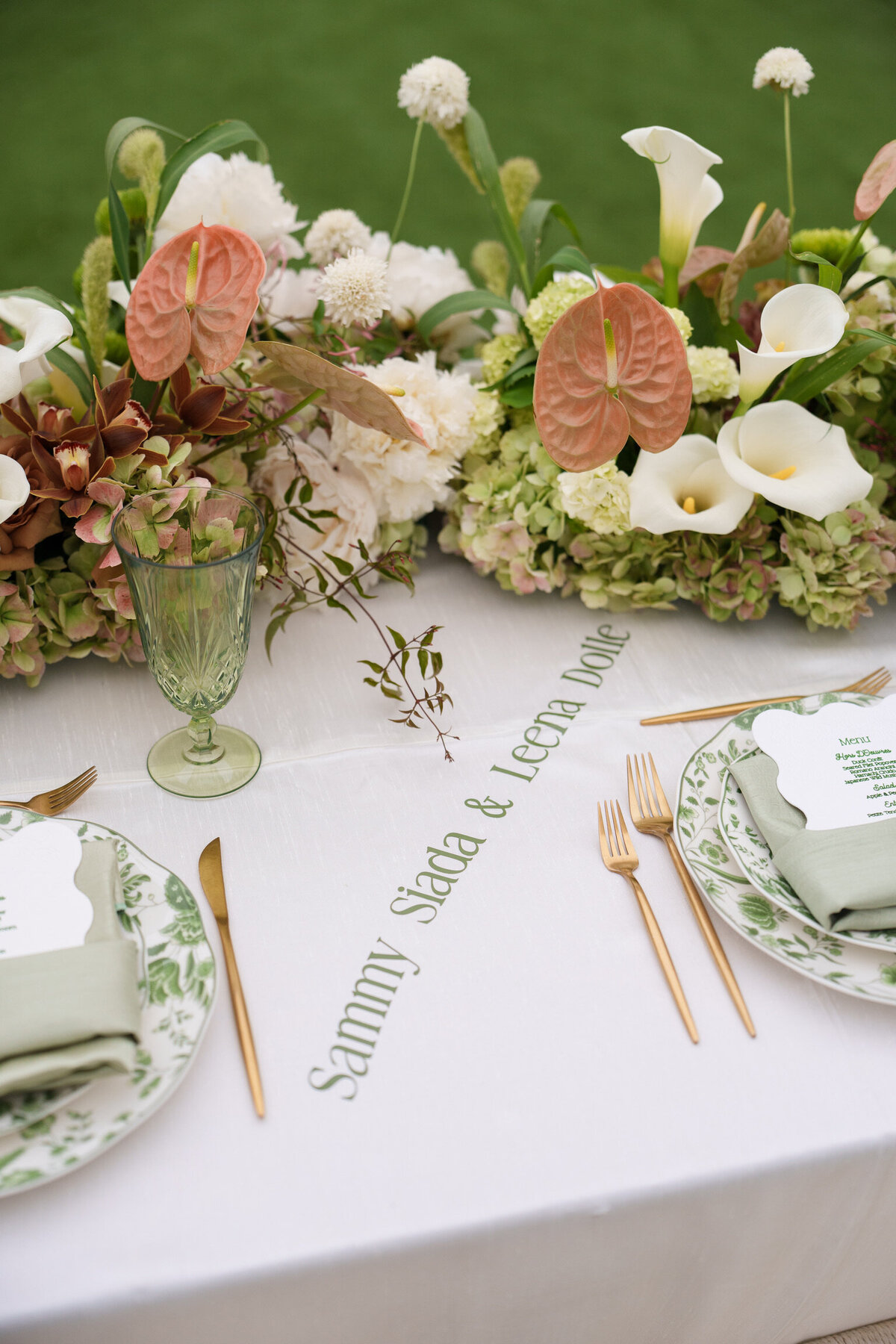 Wedding table with embroidered names, green glassware, and tropical floral arrangements.