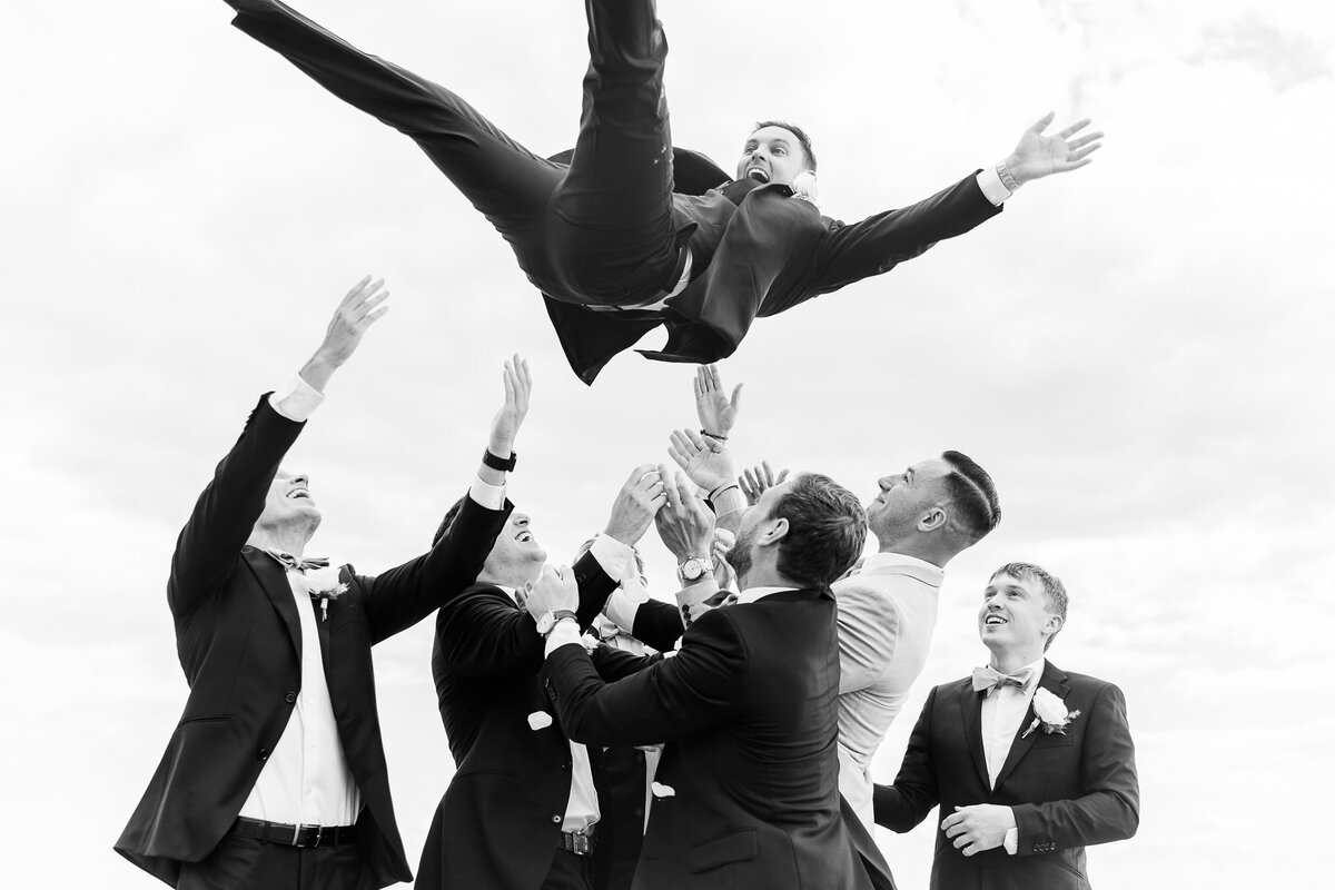 Groomsmen toss groom in the air on the shoreline of the Madison Beach Hotel. Captured by best New England wedding photographer Lia Rose Weddings.