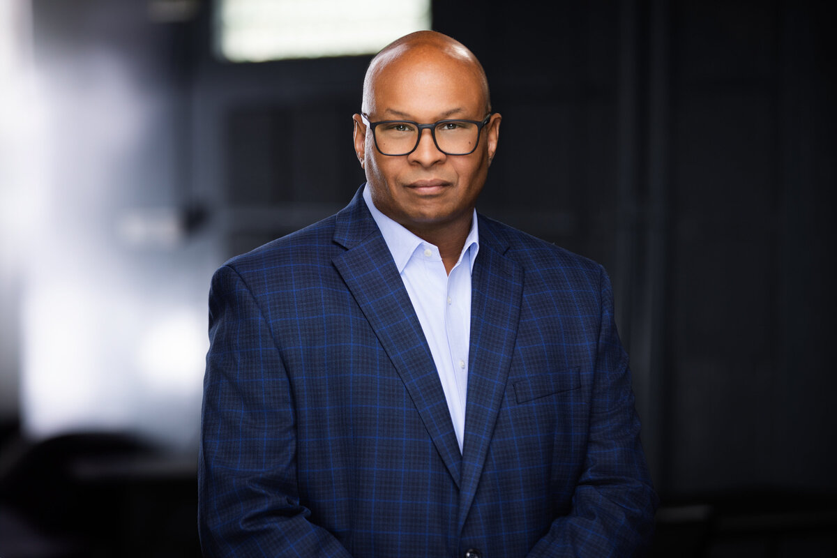 Professional headshot of an African American man with glasses and a bald head, captured in Cincinnati for personal branding. He wears a sharp blue plaid suit and a light blue shirt, exuding confidence and professionalism. Perfect for updating LinkedIn profiles and personal branding materials. Optimize your Cincinnati headshots with expert photography services for a polished, professional image.