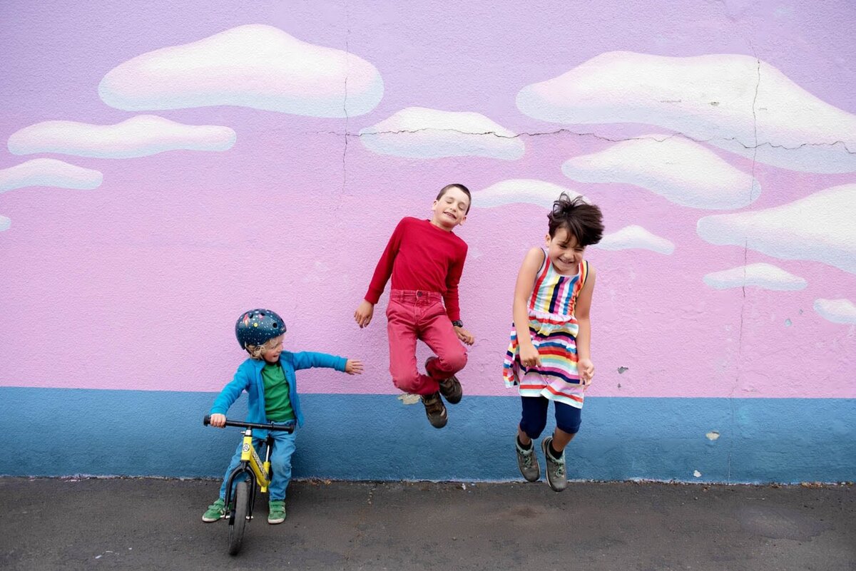 3 cute kids jumping in front of a colorful cloud mural, while one sits on bike in Portland,