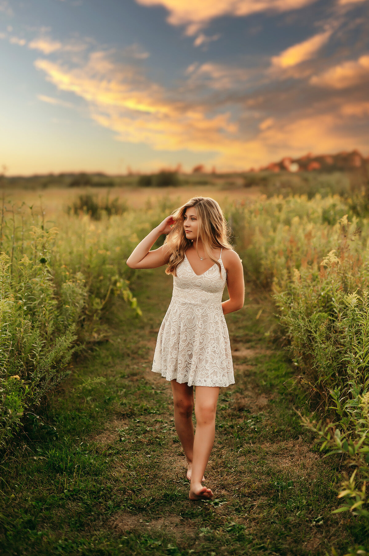 A young lady from New Berlin West High School wears a white sundress and walks barefoot through a field in Minooka Park for her senior photoshoot.