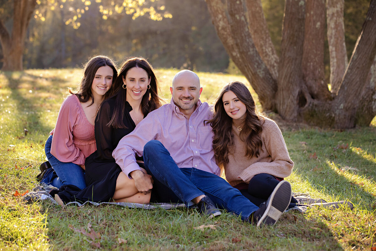 Peaceful family moment with parents and two daughters seated in grassy field surrounded by golden fall foliage at Anne Springs Close Greenway Charlotte NC