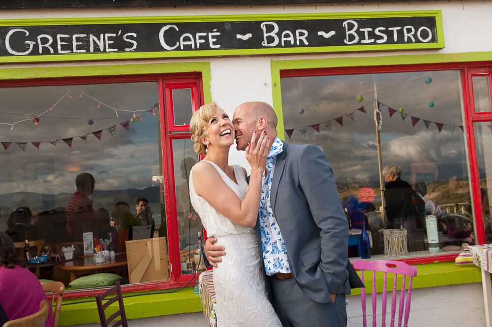 bride in v-neck column dress being kissed by groom in light grey wedding suit and floral shirt outside cafe at Inch Beach
