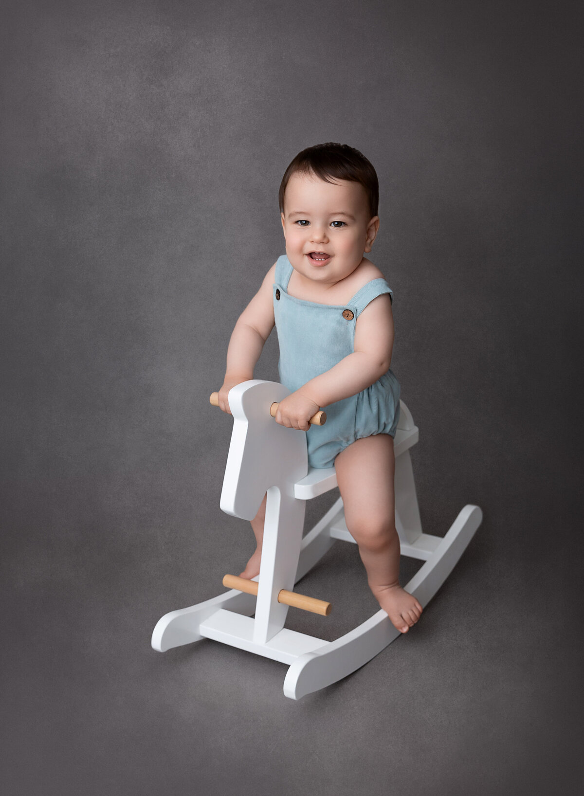 Baby boy sitting on a white rocking horse for baby milestone photoshoot. He is wearing a blue overall romper and smiling at the camera.