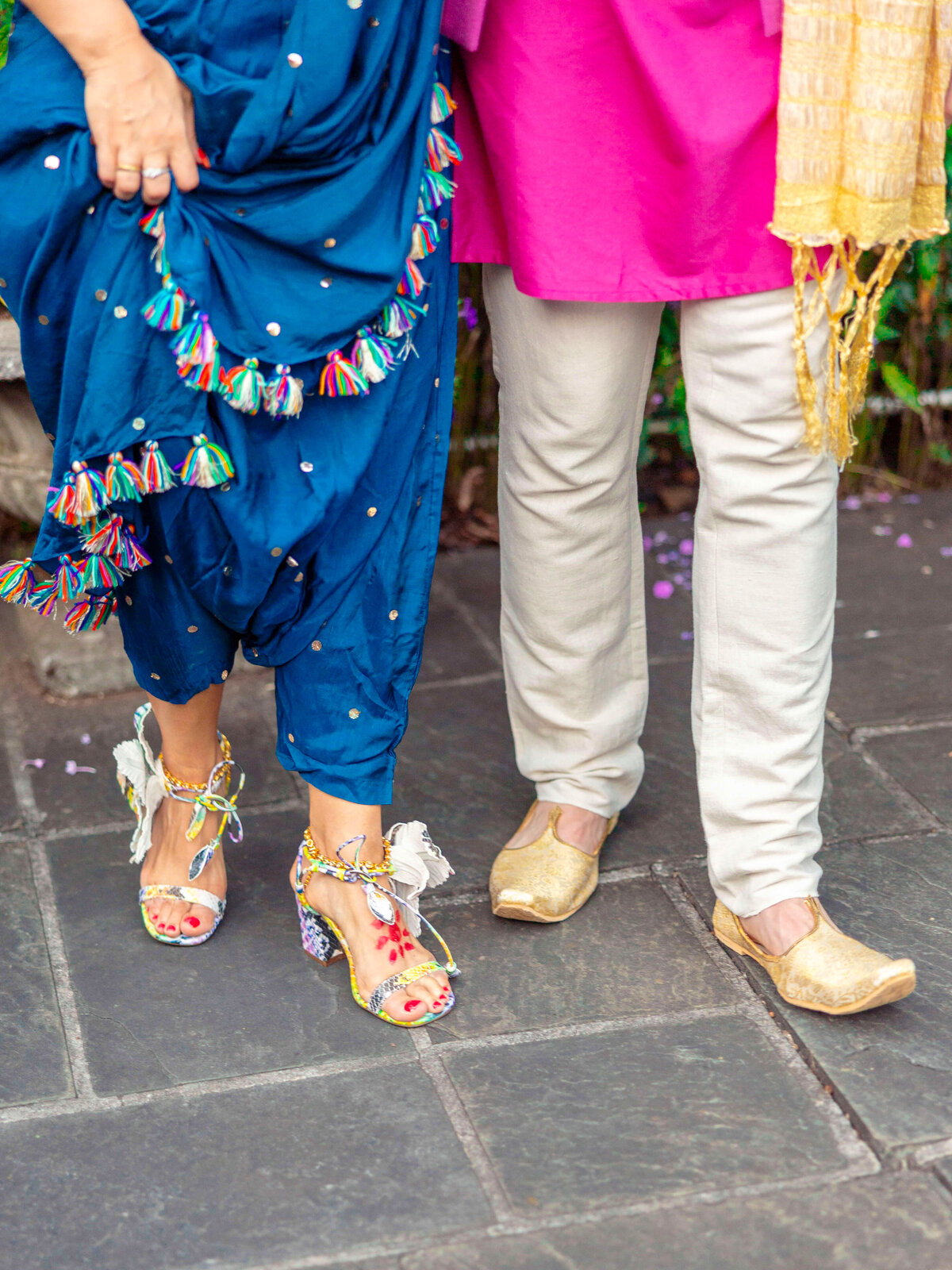 Two people stand side by side. The person on the left wears a blue dress with tassels and colorful heels. The person on the right wears a pink kurta, cream pants, and gold shoes. They are on a stone path.
