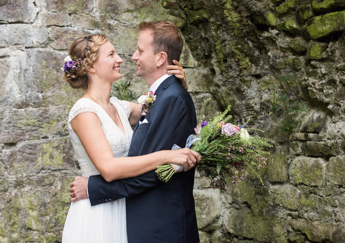 bride wearing a column style wedding dress with beaded top, holding a wild flower bouquet while embracing her groom, wearing a navy wedding suit while standing in a castle ruin