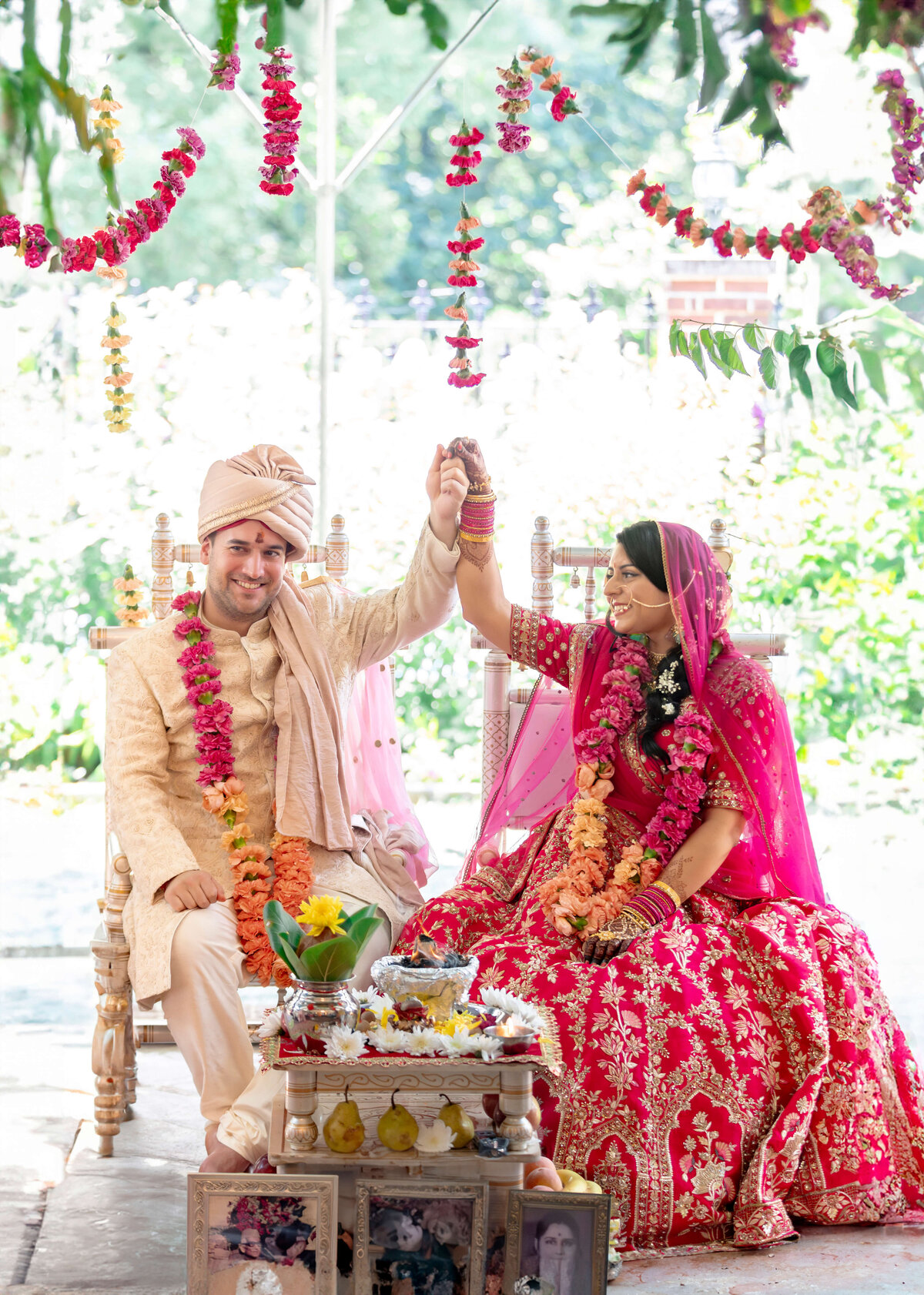 A bride and groom in traditional Indian attire smile as they hold hands under a canopy adorned with flowers. The bride wears a red lehenga, and the groom wears a beige sherwani. Floral garlands and decorations surround them.
