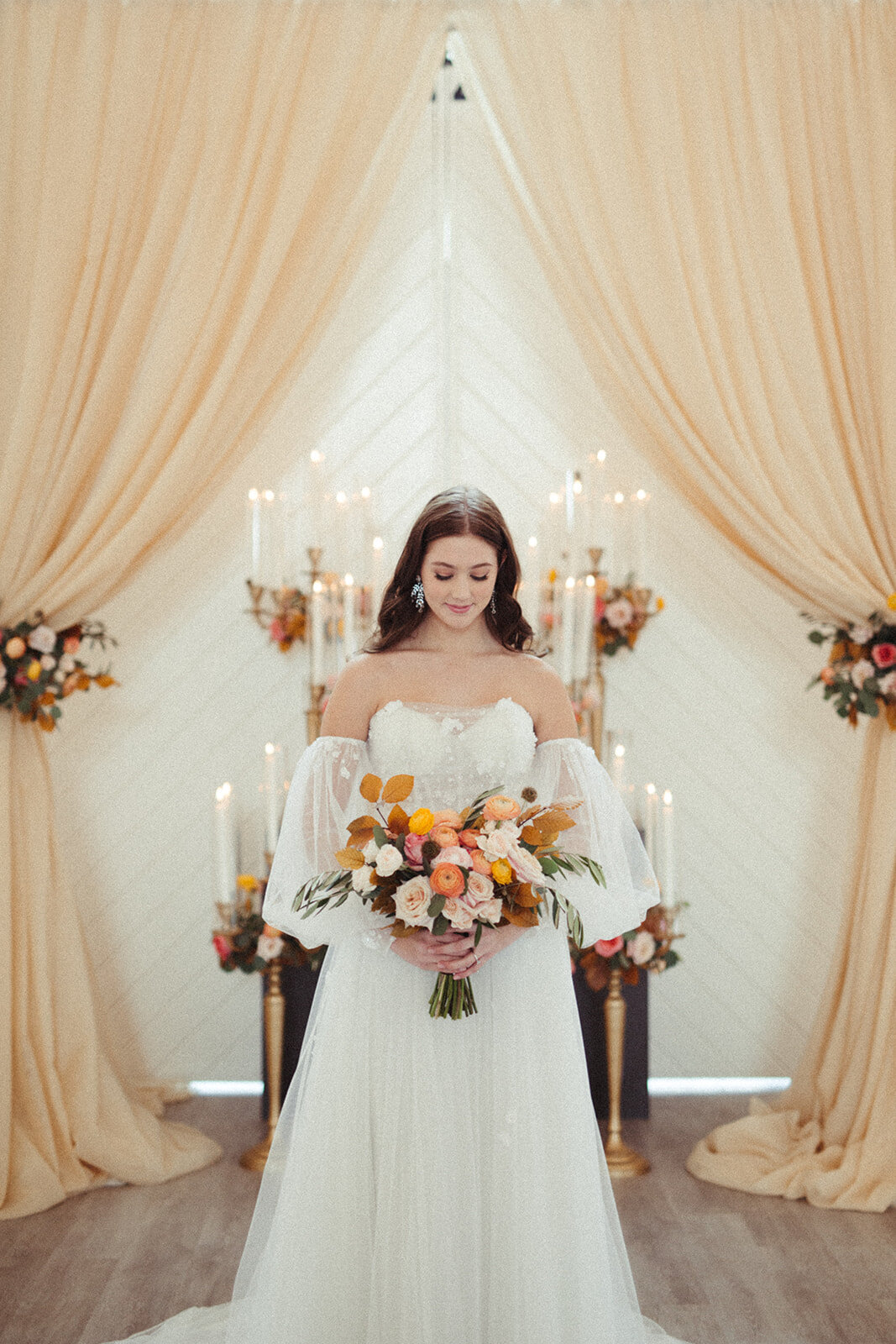 A bride wearing a white wedding gown looks down at her bouquet in a light and airy room filled with candlesticks.