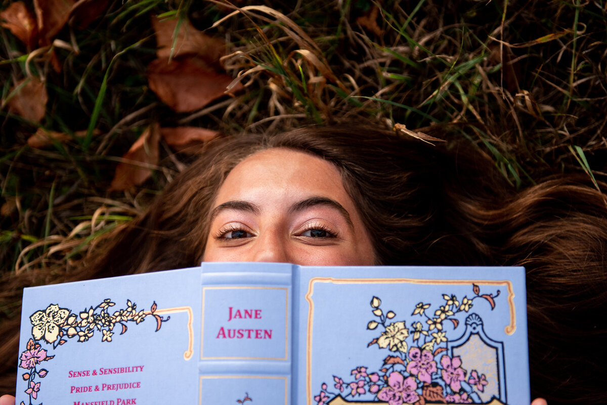 a high school girl lies in the grass with a copy of a jane austen book over her face and just her eyes peeking out