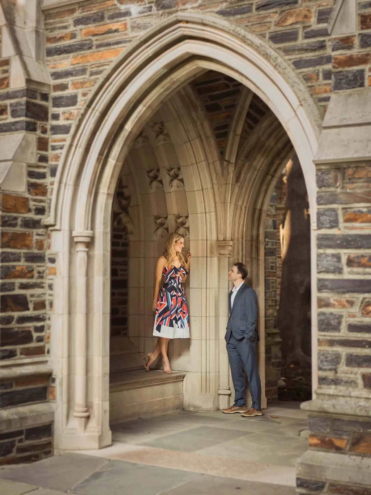 A man in a blue suit looks up at a woman in a patterned dress standing in a gothic archway