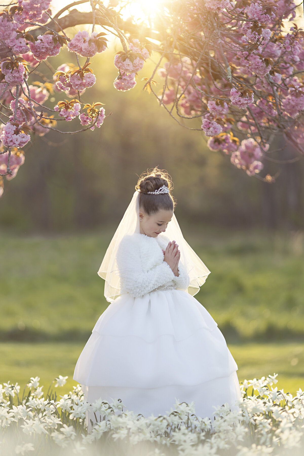 A praying young girl in a communion dress and tiara in a field of wildflowers at sunset under a pink tree