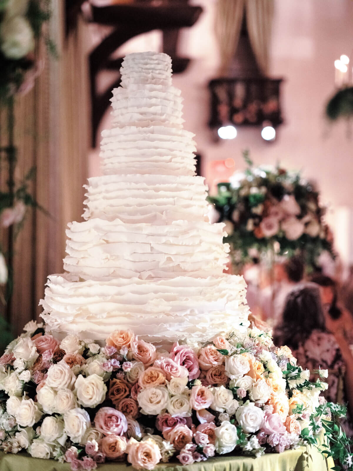 A large, white, six-layered wedding cake, with many flowers of white, pink, old rose, and peach colors at the bottom