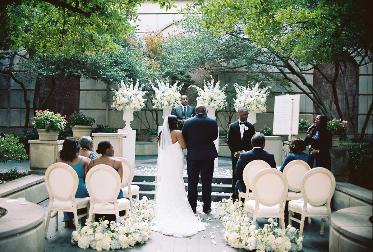 A couple stands at the altar in an outdoor garden wedding, surrounded by elegant white floral arrangements. Guests in formal attire are seated, creating a serene, joyous ambiance.
