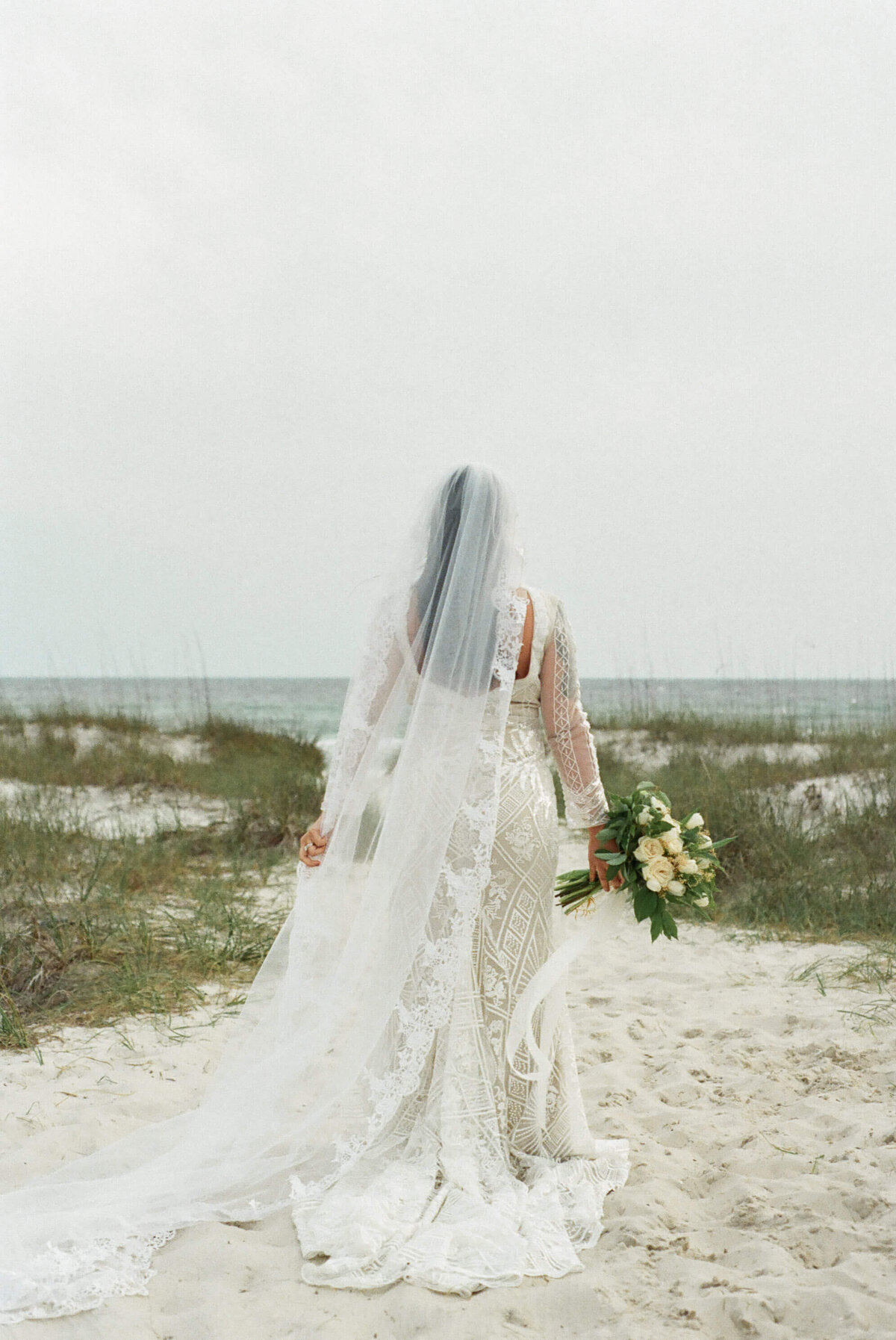 behind shot of asian bride in bridal dress and veil walks on beach path - taken by panama city fl photographer Brittney Stanley