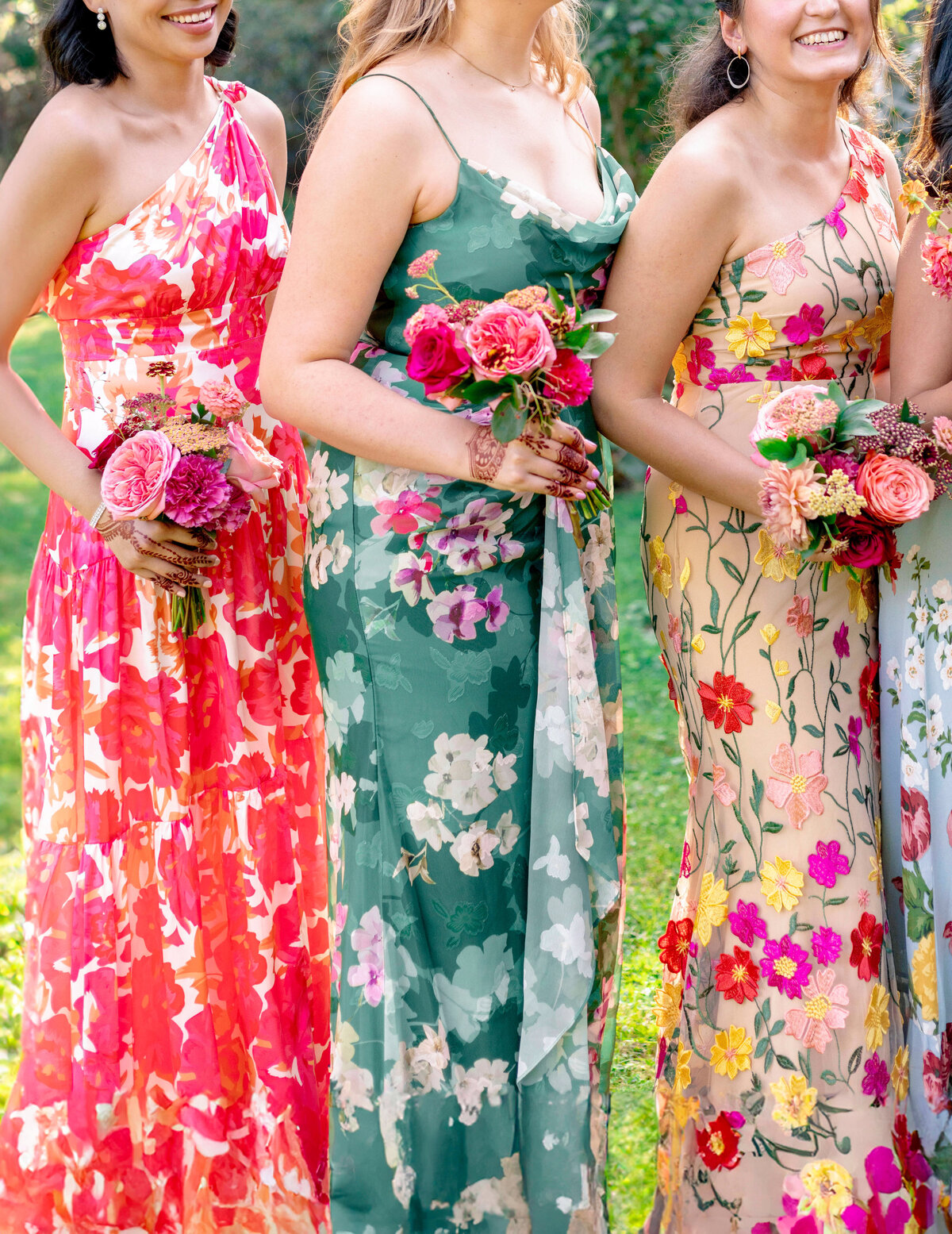 Three women standing on grass, each holding bouquets of pink roses. They wear vibrant floral dresses in shades of pink, green, and cream with various floral patterns.