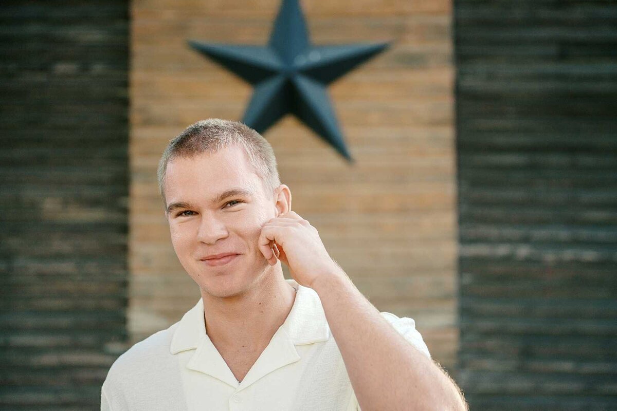 Guy senior portrait: a teen boy in a white button up shirt scratching his chin and grinning at the camera with a barn in the background