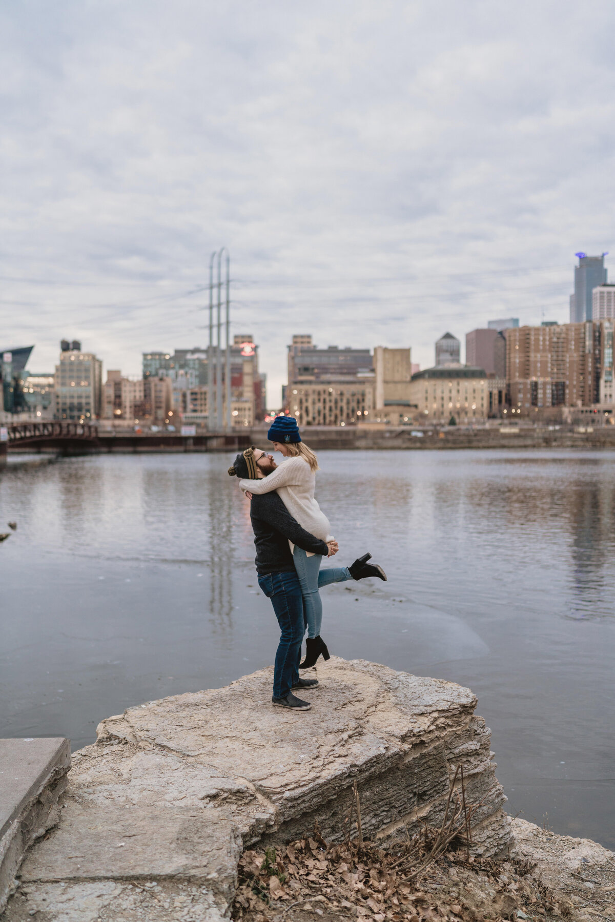 Man lifts woman on a rock in front of the Minneapolis skyline at sunrise.