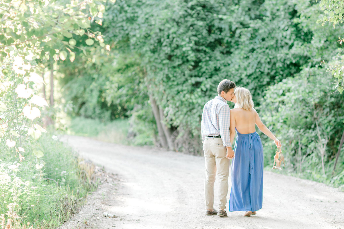couple walking hand in hand leaning in for a kiss on a gravel road lined by green trees