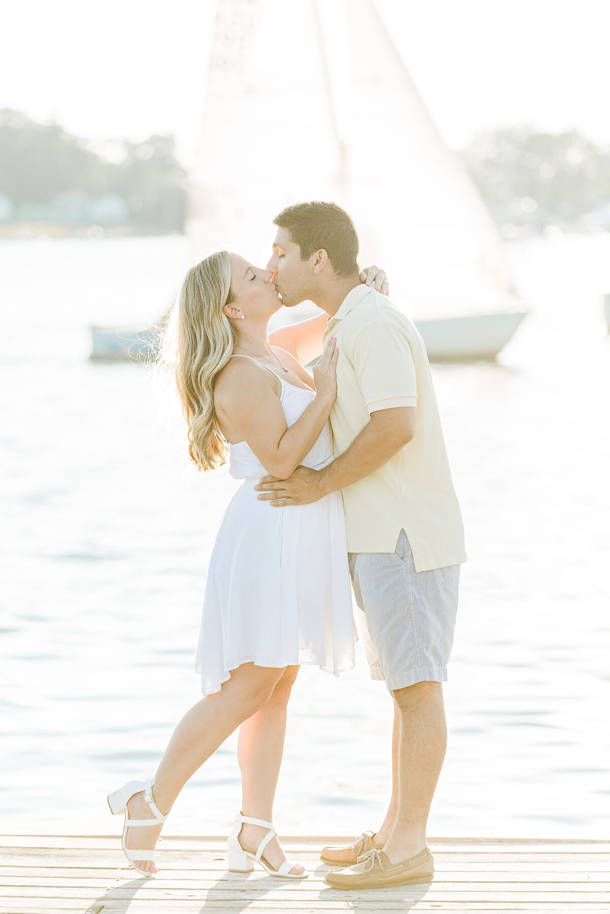 Couple share a kiss on a dock for their Bristol, RI engagement photoshoot. Their is a sailboat passing directly behind them in the background. Captured by Rhode Island Engagement Photographer Lia Rose Weddings