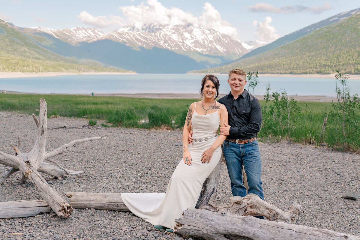 A newly married couple sitting on a log next to a lake and mountains during the midnight sun enjoying their Raleigh wedding photography session by JoLynn Photography