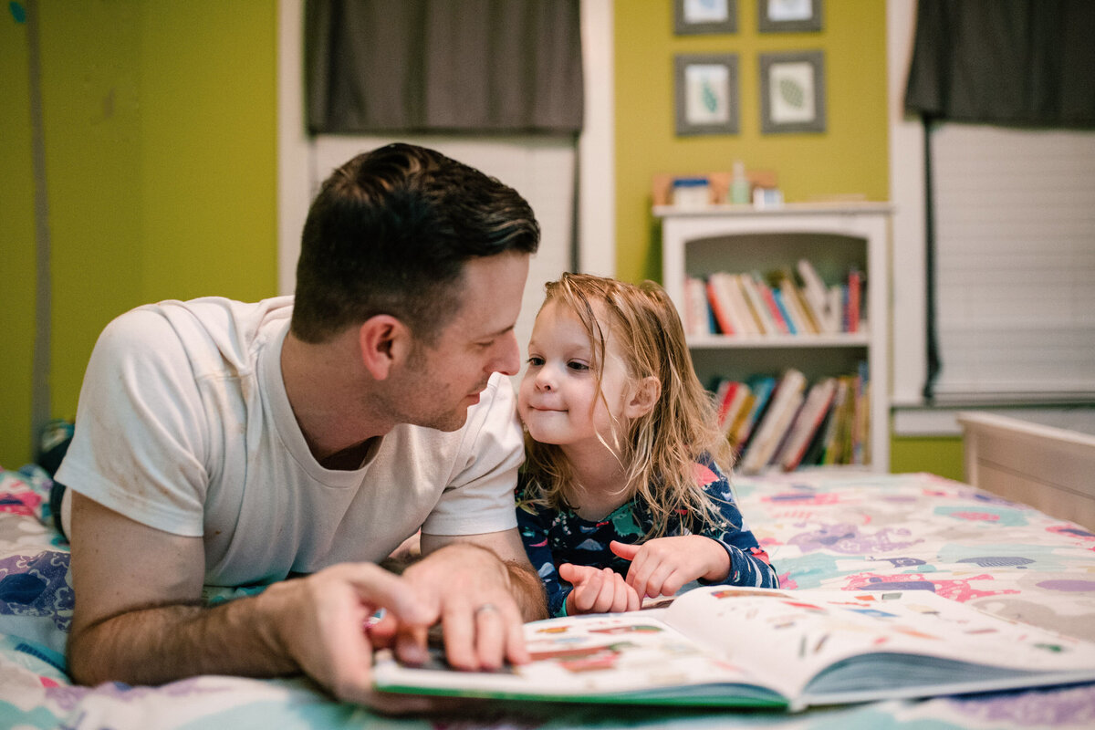 Daughter and father read a story book on a small pink bed 