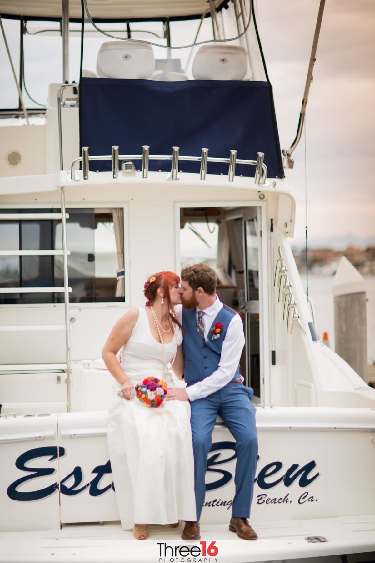 Bride and Groom sit on the edge of a boat and share a kiss