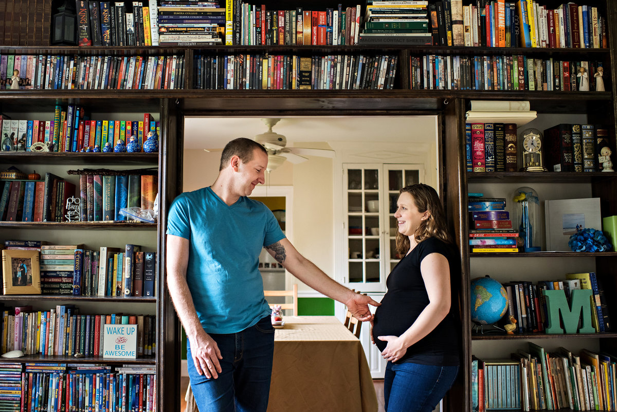 New parents to be stand under the bookshelf at their home and touch her pregnant belly.