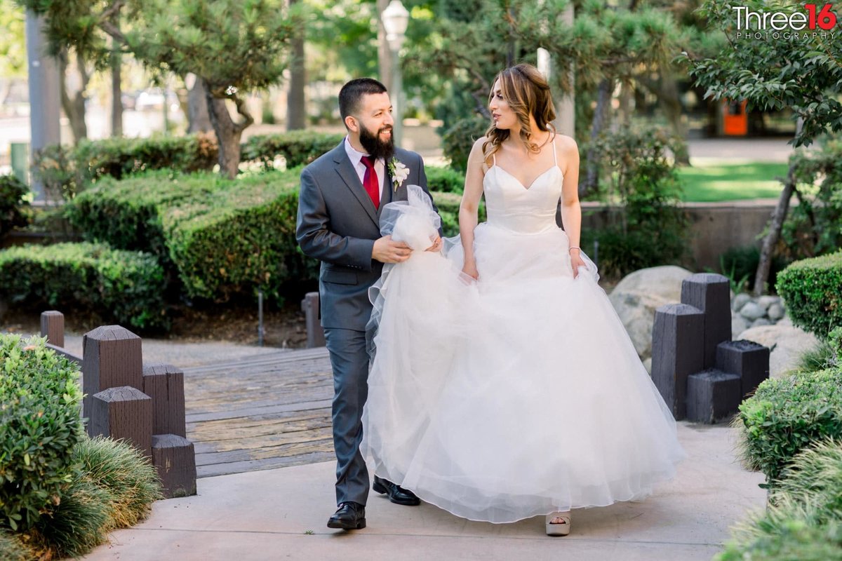 Groom carries his Bride's dress train as they walk along the grounds at the Casa Bella Wedding Venue in Anaheim