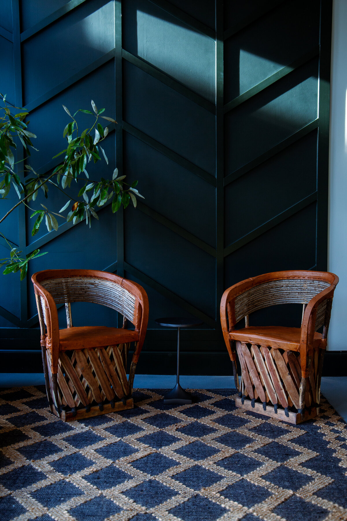 Image of two wicker and leather chairs in front of a dark green wall in a restaurants seated waiting area.