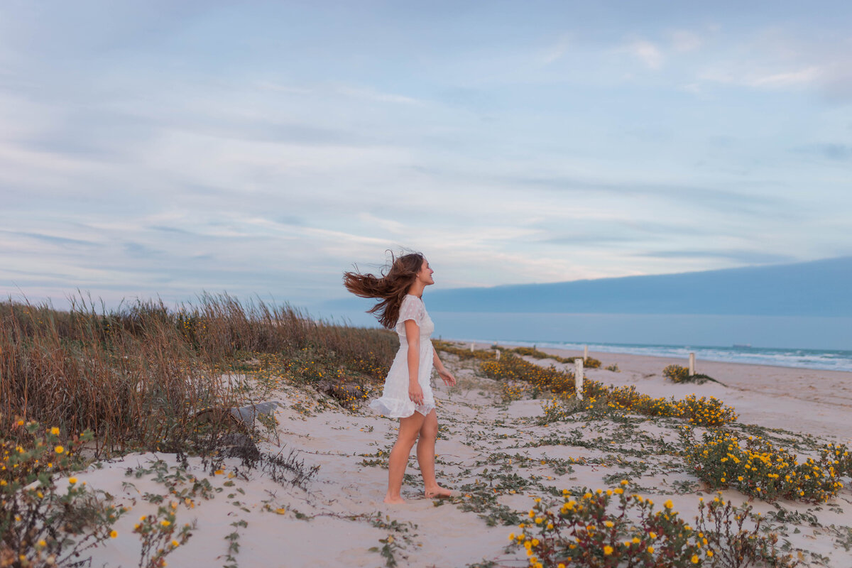 Highschool senior girl in a white dress on a sand dune at the beach on Galveston Island with wind blowing through her hair