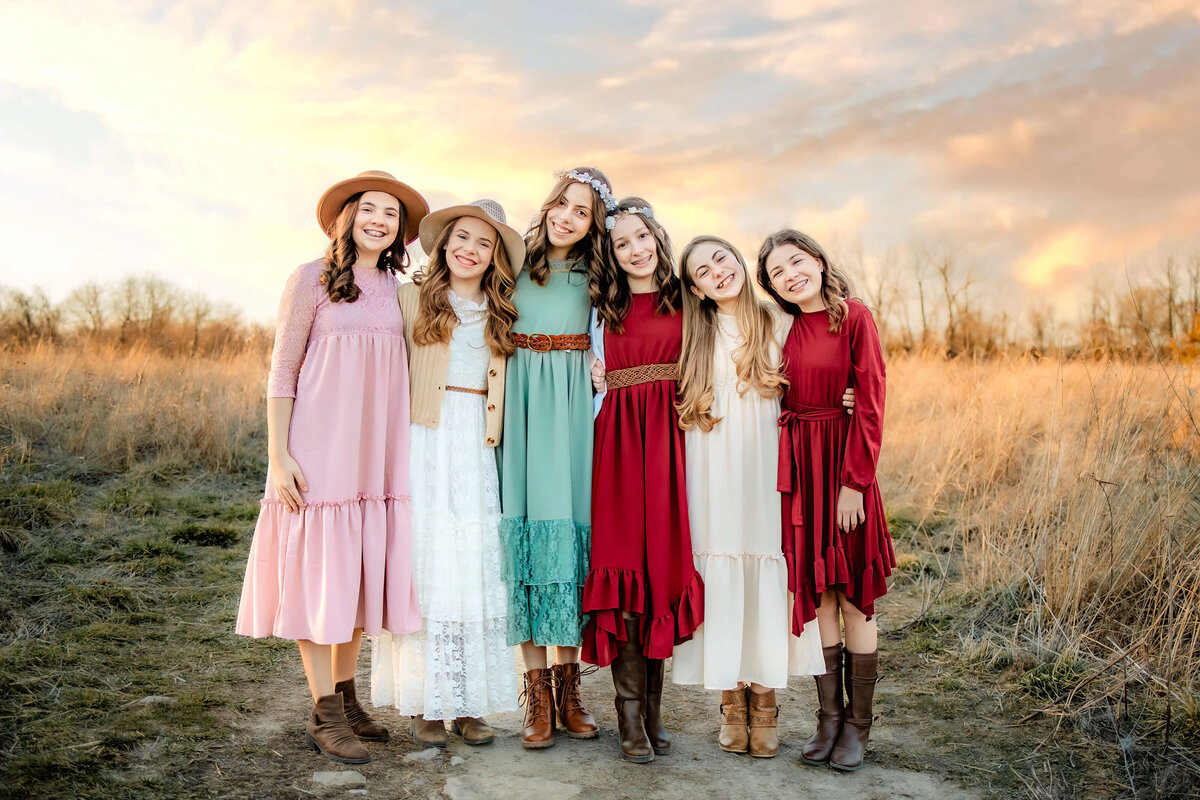 6 teenage girls in colorful dresses stading side by side with heads leaning on each other in a field at sunset