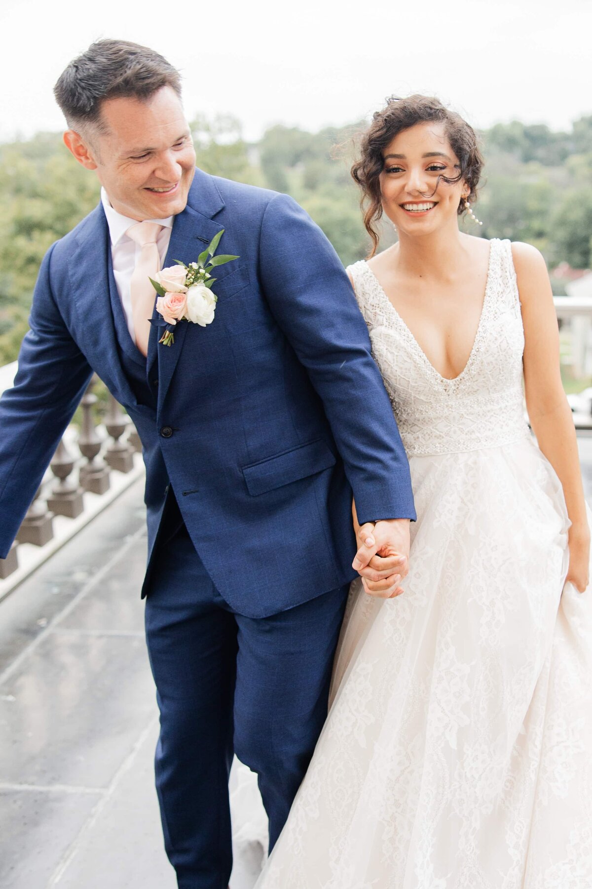 A joyful bride and groom holding hands and walking, the bride in a lace gown coordinated by a wedding planner in Des Moines, with the groom in a blue suit and pink tie.