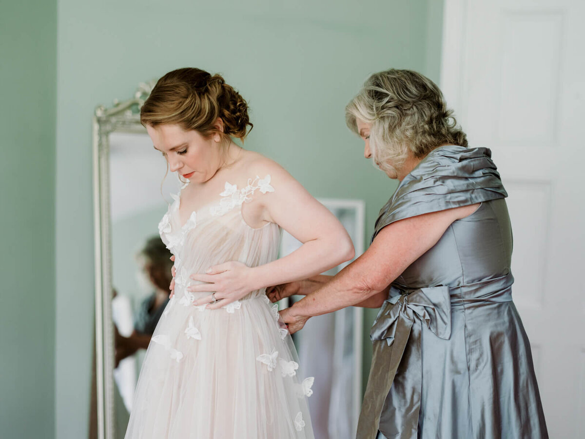 The bride is wearing her wedding dress and a woman fixing her dress from behind