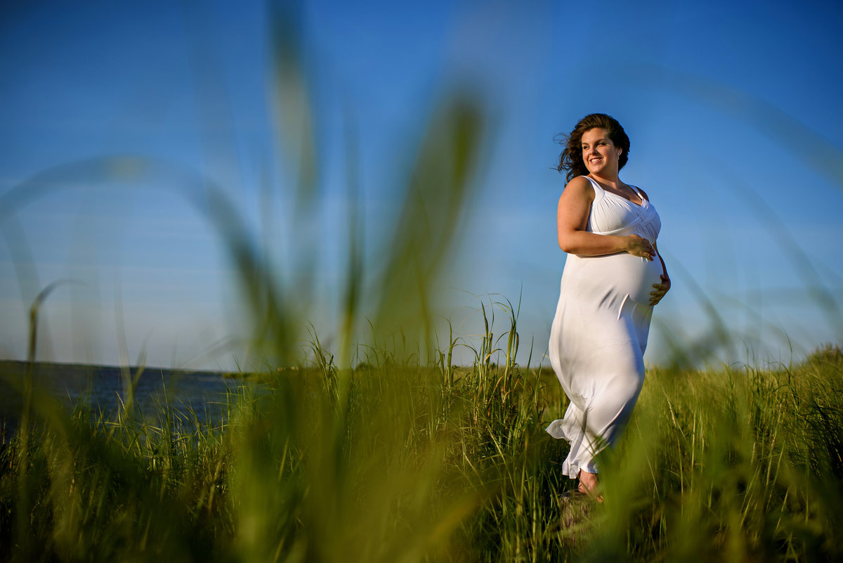 A pregnant mother at the beach stands in the tall grass and holds her pregnant belly.