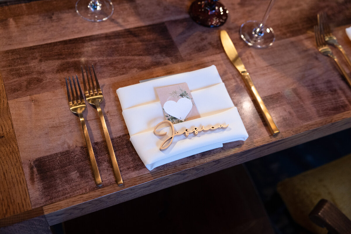 Gold silverware border napkin and wooden name sign at elegant, rustic wedding reception.
