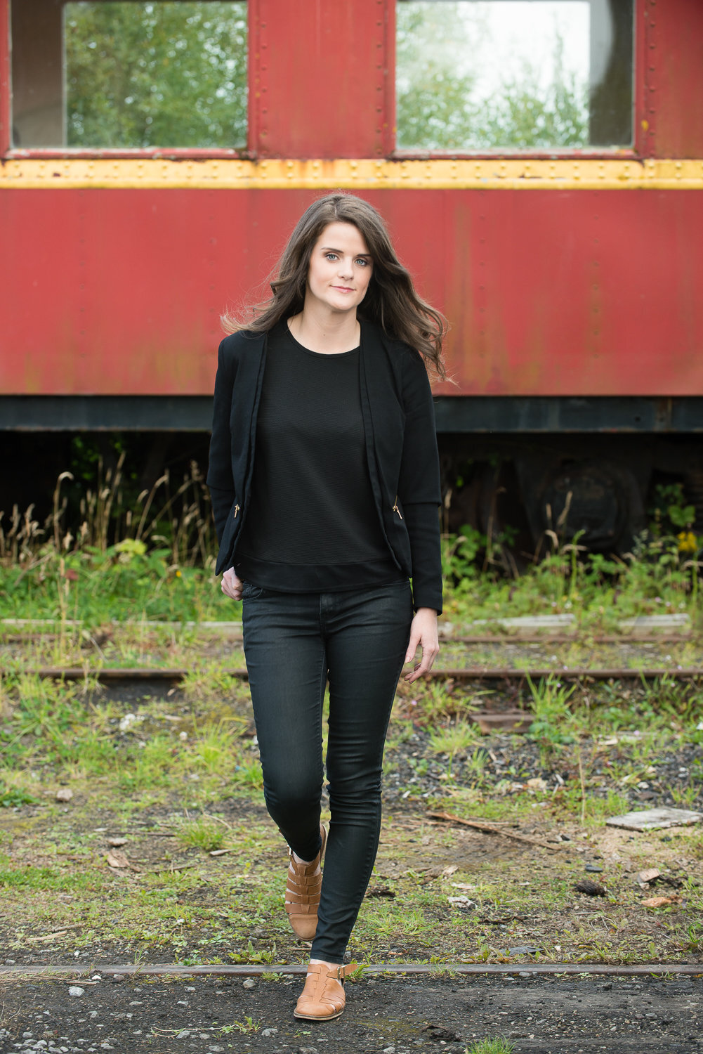 actress with long, brunette hair, in a black jeans and top, walking across train track